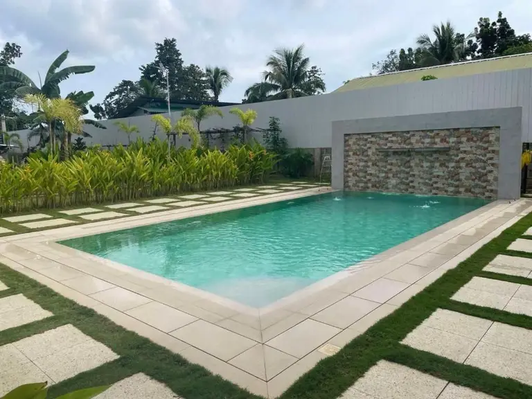Outdoor swimming pool with clear water surrounded by tiled paths and tropical plants under a cloudy sky
