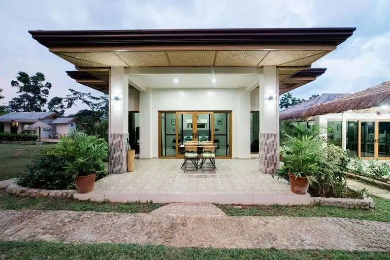 front view of a modern villa with tiled patio, wooden table and chairs, potted plants, and thatched roof cottages nearby