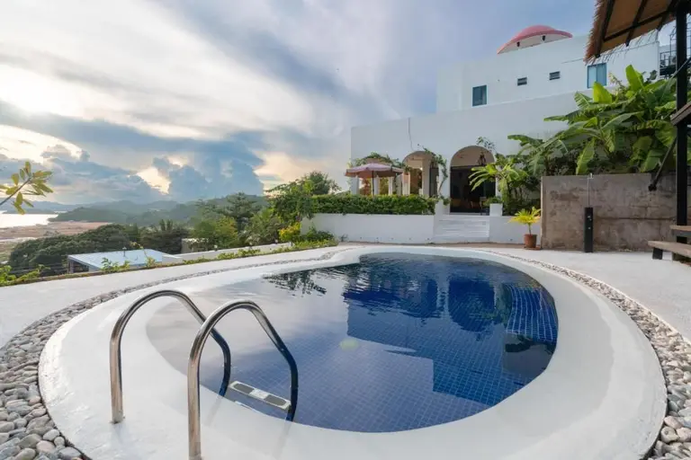 Outdoor swimming pool with metal ladder near white building and tropical plants under cloudy sky