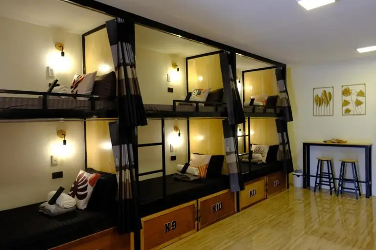 Bunk beds with black frames and curtains in a dorm room with wooden floors and stools at a counter
